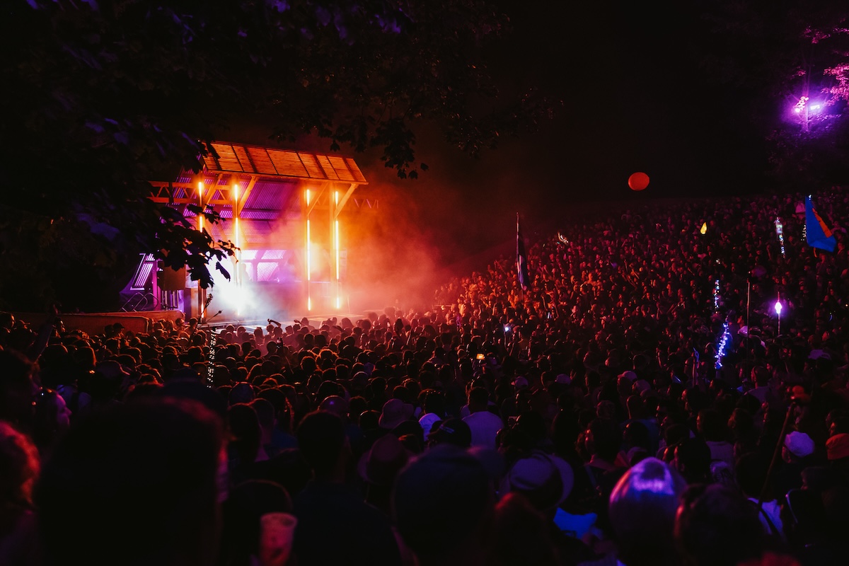 A festival stage in the dead of night beaming pink light over a teaming, dancing crowd in a bowl-shaped field