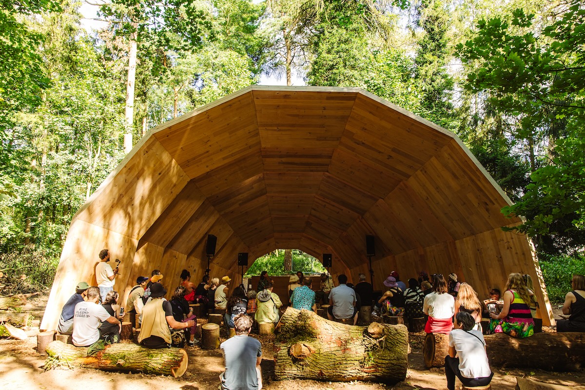 A half-tube like wooden canopy over a stage in birch woods, with people on rows of benches, picture taken from behind